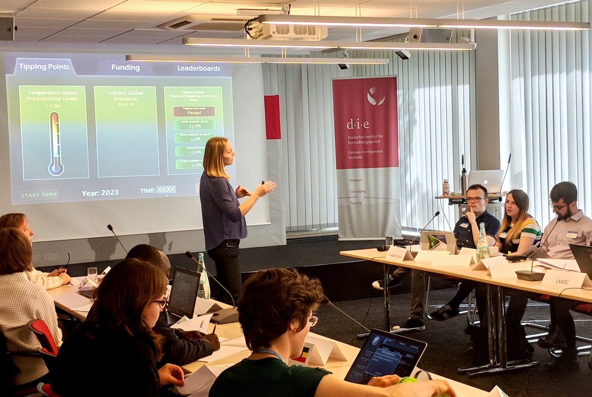Workshop participants gathered around tables, actively engaging with Gaming Climate Futures simulation materials; the room has a collaborative atmosphere with information on climate tipping points projected onto a big screen, suggesting a focus on climate policy discussion and group problem-solving